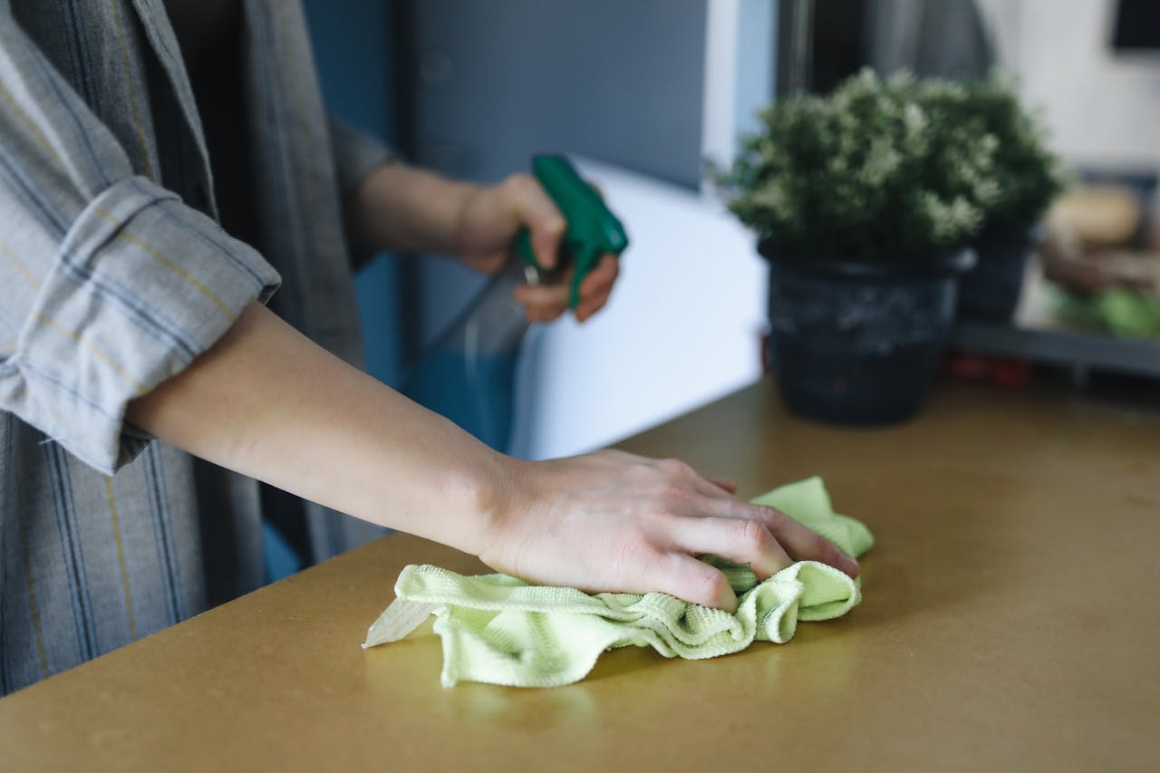 gallery-03 Close-up of a woman wiping a table with a spray bottle and cloth indoors, symbolizing effective housekeeping.